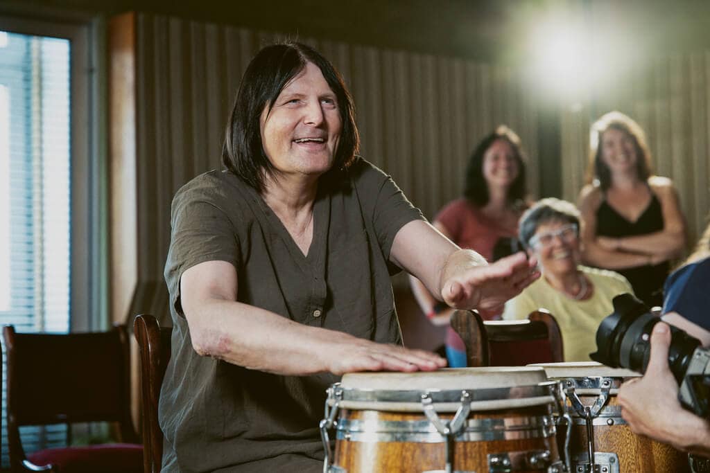 Mann im braunen Hemd spielt glücklich Bongos, Zuschauer im Hintergrund, beleuchteter Raum.