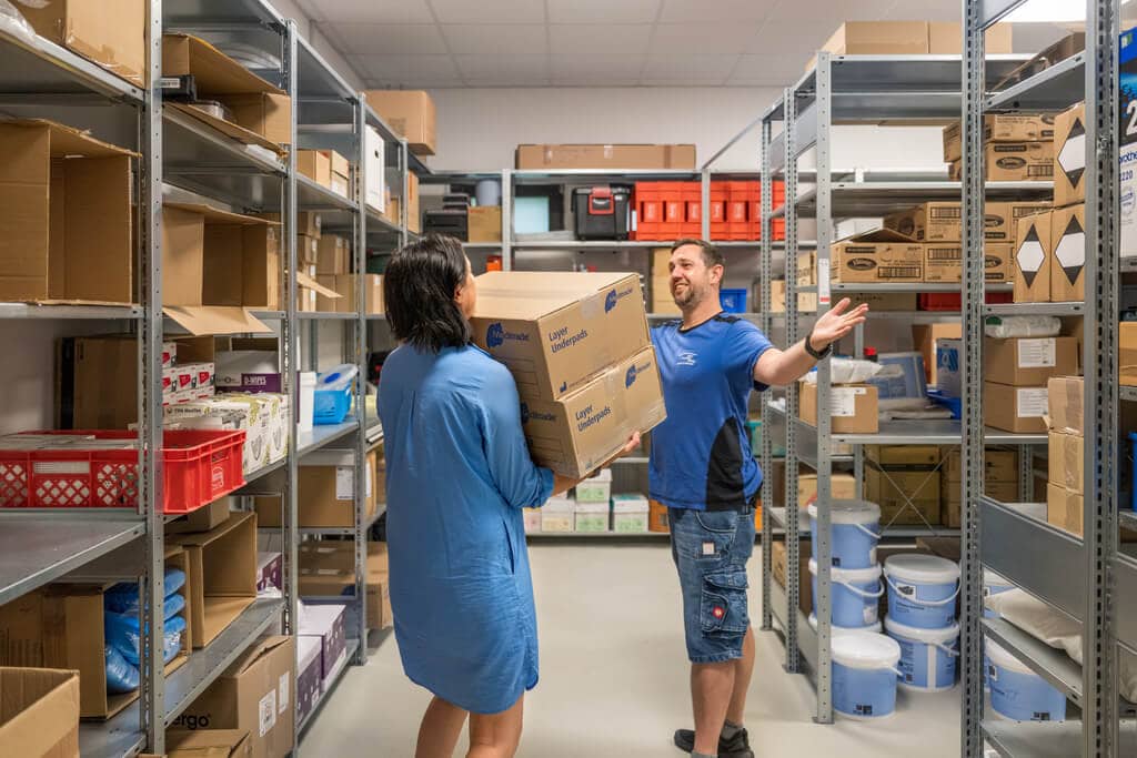 Person in blauen Shirt trägt Kartons durch Lager, ein lächelnder Kollege mit schwarzen Haaren hilft.