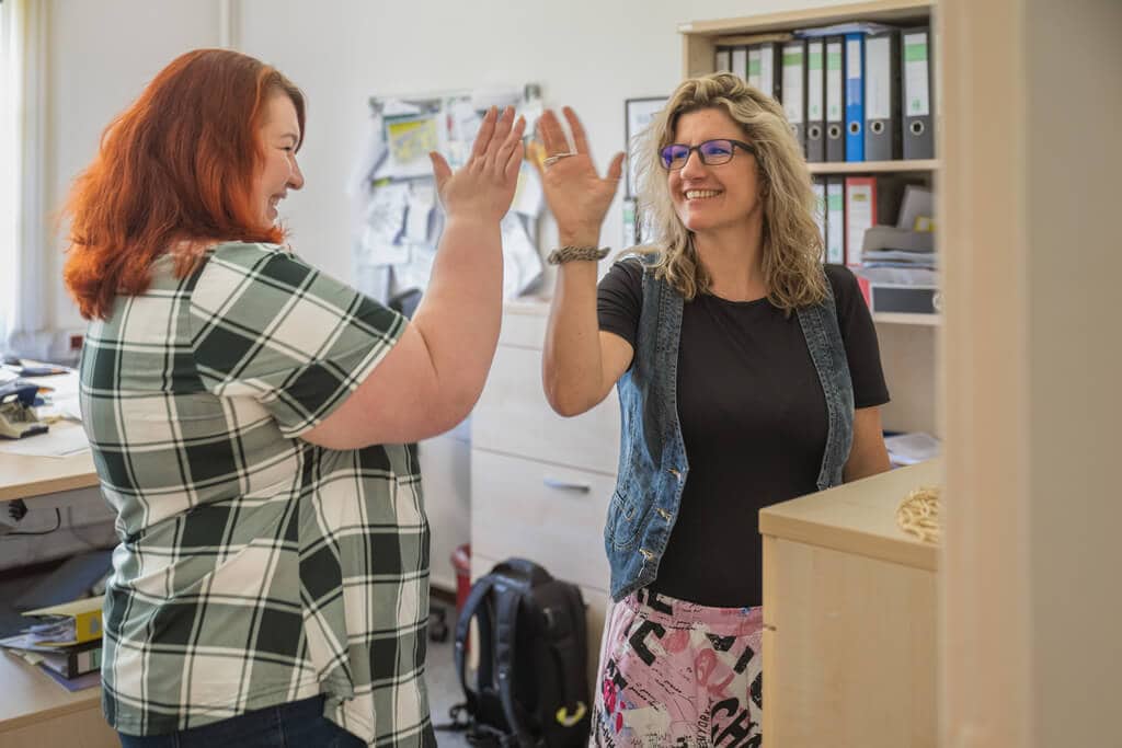 Zwei Frauen, fröhlich, im Büro, eine gibt High-Five, bunte Kleidung, Ordner im Hintergrund.