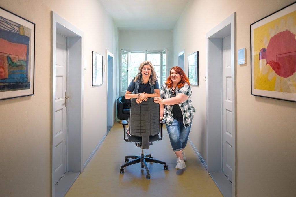 Zwei Frauen fahren mit Bürostuhl durch den Flur